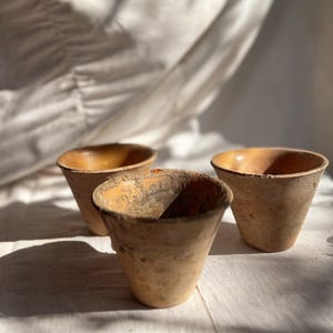 Op de afbeelding: Drie kleine, verweerde terracotta potten met een conische vorm. De potten hebben een ruw, getextureerd oppervlak en een warm, aards kleurenpalet, met tinten bruin en beige. De binnenkant van de potten heeft een lichtere, gouden tint.