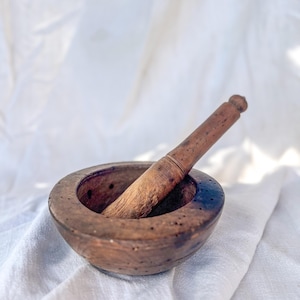 May include: A wooden mortar and pestle, with a dark brown finish. The mortar is a round bowl with small holes around the rim. The pestle is a long, cylindrical piece of wood with a rounded end. The set is displayed on a white cloth.