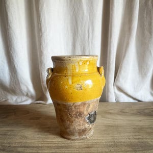 May include: A ceramic vessel featuring a bright yellow glazed upper section with visible drips and a natural, unglazed terracotta base. The pot has two small side handles and a weathered, textured surface with a dark circular mark on the lower body.