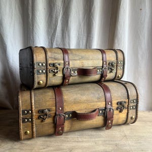 Może przedstawiać: Dwie drewniane walizki w stylu vintage ułożone jedna na drugiej. Mają jasnobrązowe wykończenie z ciemnobrązowymi skórzanymi paskami i uchwytami. Metalowe zapięcia i nity dodają uroku antycznemu wyglądowi. Walizki leżą na drewnianej powierzchni.
