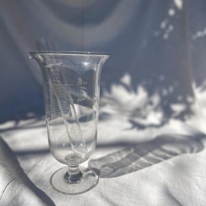 May include: A clear glass beaker with a flared top and a slender, graduated pipette inside. The beaker has measurement markings and sits on a round base. The image is lit with natural light, casting shadows.