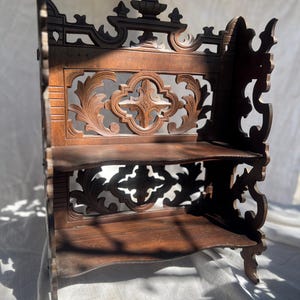 Puede incluir: Estantería de esquina de madera oscura y antigua con tallas ornamentadas. La estantería tiene dos niveles y un respaldo decorativo con diseños florales y geométricos calados. La madera tiene un tono rico y cálido.