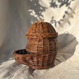 May include: A handcrafted, brown wicker basket with a domed lid and a small, rectangular tray attached. The basket is made of woven twigs and has a natural, rustic appearance. The lid has a small loop for easy opening.