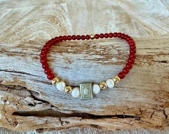 May include: A red beaded bracelet with gold beads and a white, square charm with a geometric design.