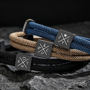 Peut inclure: Trois bracelets en corde noire, beige et bleu marine, chacun avec un fermoir carré en métal gris foncé. Les fermoirs présentent un motif de croix blanche avec des lettres. Les bracelets sont empilés sur une surface rocheuse texturée gris foncé.