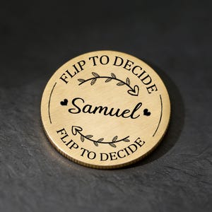 May include: A gold-colored decision coin with the text "Flip to Decide" around the edge and the name "Samuel" in the center. The coin has a decorative leaf design and small hearts. The coin is on a dark, textured surface.