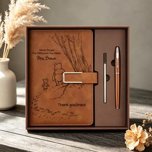 May include: A tan leather-like journal with a Winnie the Pooh illustration and the text "Never Forget The Difference You Make Mrs. Brown" and "Thank you Grace," featuring a metal clasp, paired with a matching pen in a brown presentation box.