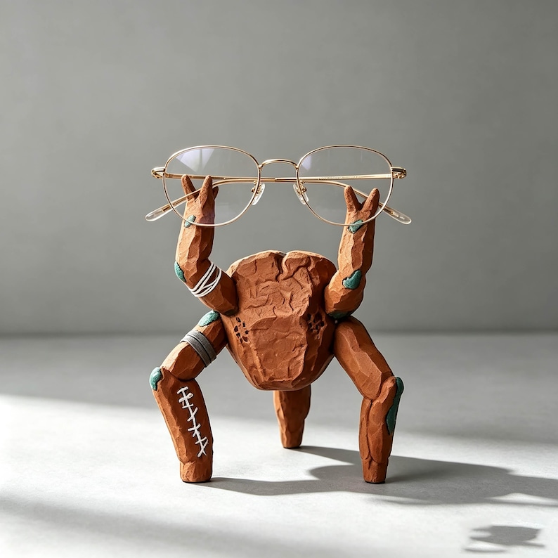 May include: A brown, textured, carved-style creature figurine with multiple limbs holding a pair of thin, gold-rimmed eyeglasses. The figure features white stitched details on one leg, white bands on an arm, and small teal accents at the joints.
