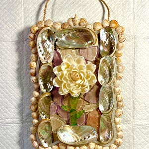 May include: A rectangular wall hanging decorated with seashells, abalone shells, and a central flower design. The frame is lined with small, round shells. The flower is cream-colored with pearl accents, and the stem is green. A rope loop is attached for hanging.