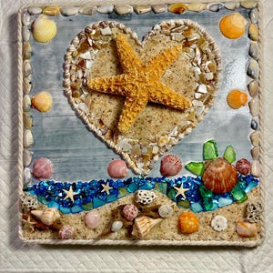 Pode incluir: Uma obra de arte quadrada com tema de praia, apresentando um design em forma de coração preenchido com areia e uma estrela do mar. A obra é adornada com várias conchas, vidro do mar e uma borda de corda. A paleta de cores inclui tons de bege, azul, laranja e branco.
