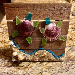 Pode incluir: Uma caixa de madeira decorada com duas tartarugas de conchas. As tartarugas têm conchas roxas, detalhes de barbatanas de vidro verde e cabeças de vidro azul. A base apresenta areia, conchas e pedras azuis e brancas, criando um tema costeiro.