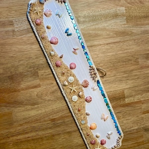Pode incluir: Placa retangular de madeira branca decorada com conchas, estrelas do mar e areia. As bordas são adornadas com corda e pequenas conchas. Uma linha de contas iridescentes azuis e verdes percorre um lado. Decoração com tema de praia.