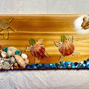 Pode incluir: Placa de madeira com tema de praia. Apresenta três tartarugas de concha, uma estrela do mar e uma borboleta de vidro. A base é decorada com conchas e pedaços de vidro azul.