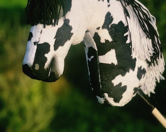 Puede incluir: Un caballo de juguete hecho a mano con un patrón de manchas blancas y negras. La crin y el flequillo del caballo son negros, y tiene un mango de madera. El fondo es verde borroso.