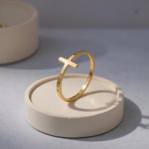 May include: A gold-colored ring featuring a cross design. The ring is displayed on a round, off-white surface. The cross is positioned on the side of the ring, creating a unique and modern look. The ring's band is thin and simple.