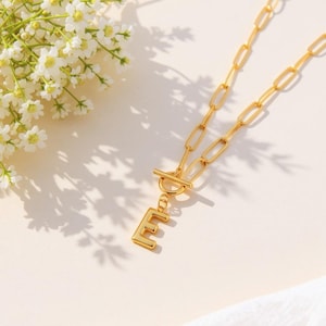 May include: A gold-colored necklace with a large link chain and a toggle clasp. The pendant is the letter "E". White flowers are in the upper left corner. The necklace is on a cream-colored surface.