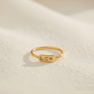 May include: A gold ring with a rectangular plate bearing the letters "L", a heart symbol, and "K". The ring sits on a soft, off-white surface. This delicate piece of jewellery is a simple yet elegant design.