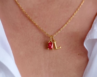 Könnte beinhalten: Eine goldfarbene Halskette mit feiner Kette. Der Anhänger zeigt einen rechteckigen, roten Edelstein und den goldenen Buchstaben "L". Die Halskette wird auf einem weißen Hemd getragen.