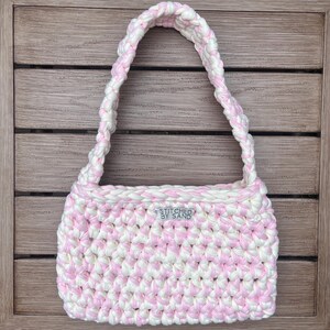 Pu&ograve; includere: Borsa a mano all'uncinetto a righe rosa e bianche. La borsa ha una forma rettangolare, un manico intrecciato e l'etichetta "STITCHED BY SAND". Fatta a mano.