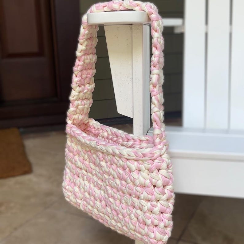 Pu&ograve; includere: Borsa a tracolla all'uncinetto rosa e bianca con tracolla intrecciata. La borsa presenta un motivo intrecciato e una forma rettangolare. La borsa &egrave; realizzata con filato spesso. La borsa &egrave; appesa a un palo di legno bianco.
