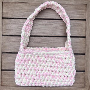 Pu&ograve; includere: Borsa all'uncinetto fatta a mano con un motivo rosa e bianco. La borsa ha un corpo rettangolare e un manico abbinato. Le cuciture all'uncinetto sono visibili, creando una superficie strutturata. La borsa &egrave; progettata per trasportare oggetti.