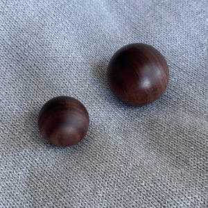 Peut inclure: Deux sphères en bois brun foncé et lisse de tailles différentes reposent sur un tissu gris texturé. La plus grande sphère mesure environ 5 cm de diamètre, la plus petite environ 2,5 cm.