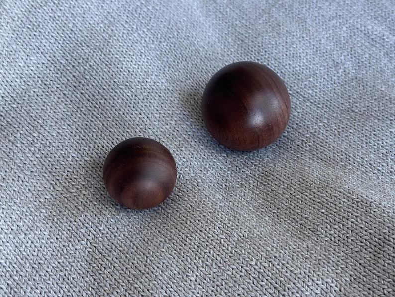 May include: Two smooth, dark brown wooden spheres of different sizes sit on grey, textured fabric. The larger sphere is approx. 5 cm in diameter, the smaller one is about 2.5 cm. The spheres have a polished finish.