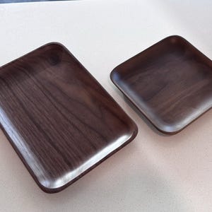 May include: Two dark brown wooden trays. The trays are rectangular with rounded corners and edges. The wood grain is visible on the surface. These trays are likely for serving or display purposes, and are made of wood.