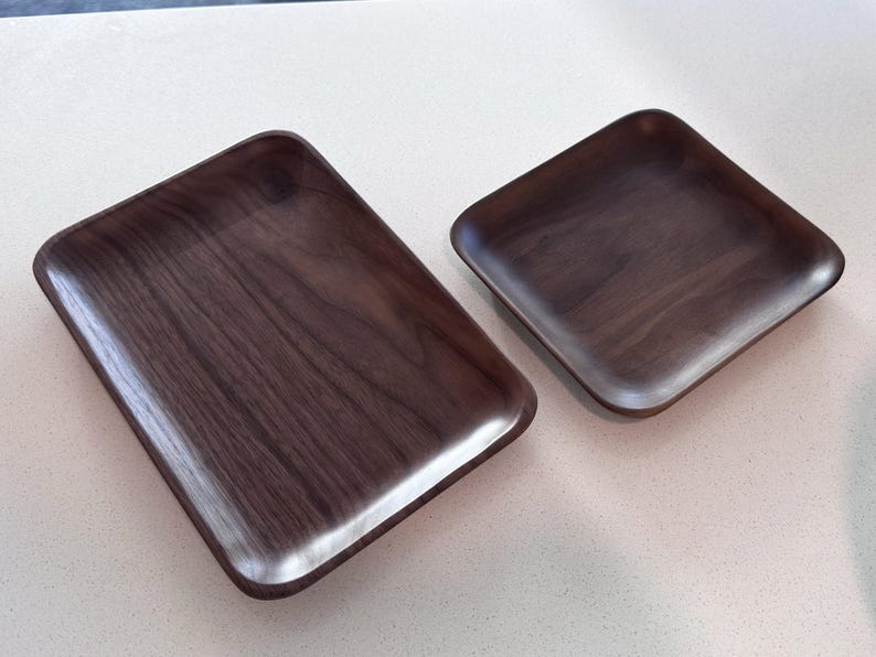 May include: Two wooden trays with a dark brown finish. The trays have a rectangular shape with rounded corners and edges. The wood grain is visible on the surface of the trays. The trays are likely used for serving or display.