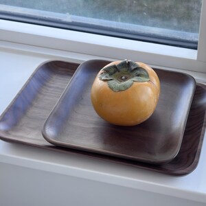 May include: Two dark brown wooden trays, one stacked on the other, with rounded corners. A ripe orange persimmon fruit with a green calyx sits on the top tray. The trays are on a white windowsill.