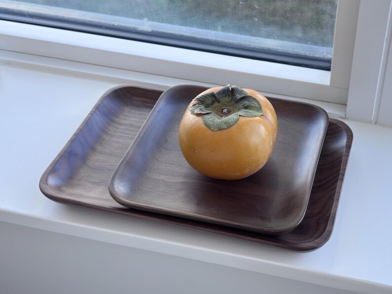 May include: Two dark brown wooden trays, one stacked on the other, with rounded corners. A ripe orange persimmon fruit with a green calyx sits on the top tray. The trays are on a white windowsill.