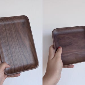 May include: Two images of a rectangular wooden tray with rounded corners. The tray is a dark brown color with visible wood grain. One image shows the tray held in a hand, and the other shows the tray on its own.