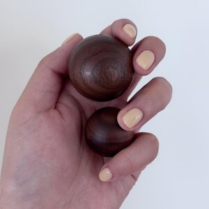 May include: Two smooth, dark brown wooden spheres held in a hand with light-coloured nail polish. The spheres have a polished finish, and the wood grain is visible. The background is plain white.