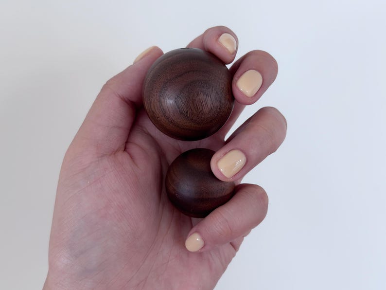 May include: Two smooth, dark brown wooden spheres held in a hand with light-coloured nail polish. The spheres have a polished finish, and the wood grain is visible. The background is plain white.