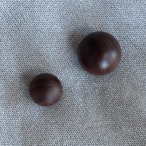 May include: Two dark brown wooden spheres of different sizes sit on a grey, textured surface. The larger sphere is approximately 5 cm in diameter, while the smaller one is about 2.5 cm across. The spheres have a smooth, polished finish.