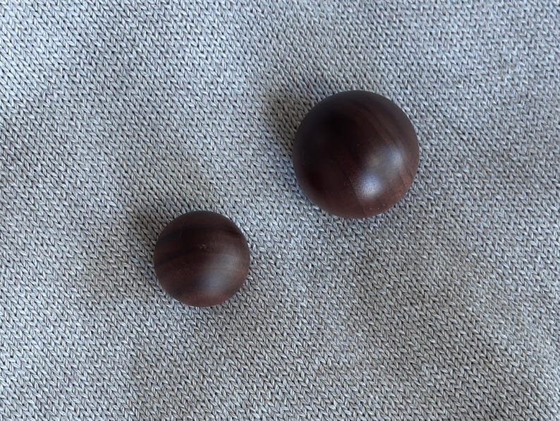 May include: Two dark brown wooden spheres of different sizes sit on a grey, textured surface. The larger sphere is approximately 5 cm in diameter, while the smaller one is about 2.5 cm across. The spheres have a smooth, polished finish.