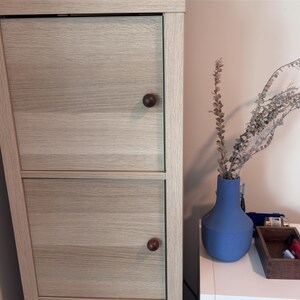 May include: A light-coloured wooden cabinet with two doors and round brown knobs. A blue vase holds dried plants. A small wooden box and other items sit on a white surface.