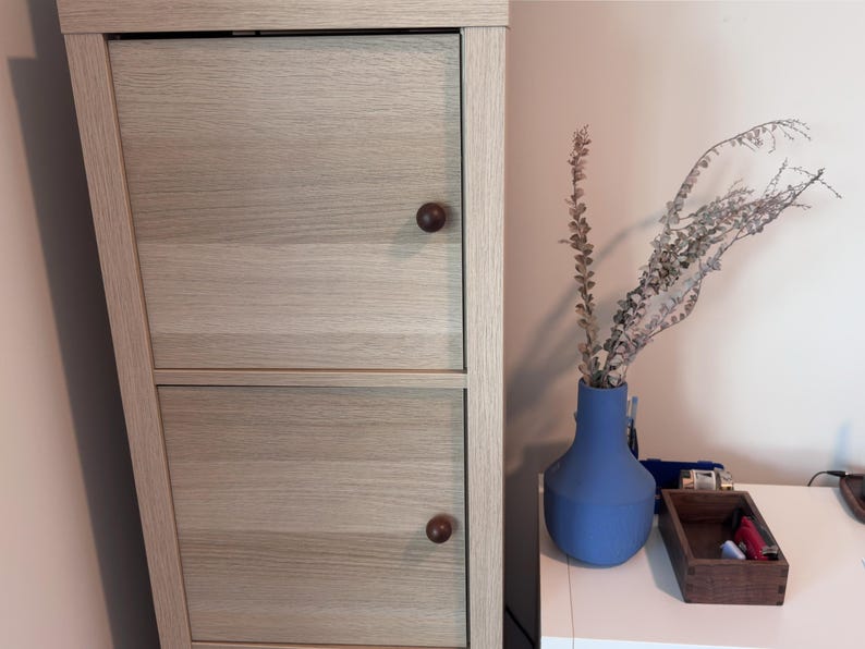 May include: A light-coloured wooden cabinet with two doors and round brown knobs. A blue vase holds dried plants. A small wooden box and other items sit on a white surface.