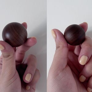 May include: Two dark brown wooden spheres, each held in a hand with light yellow nail polish. The spheres have a smooth, polished finish and a visible wood grain pattern. The background is a plain white wall.