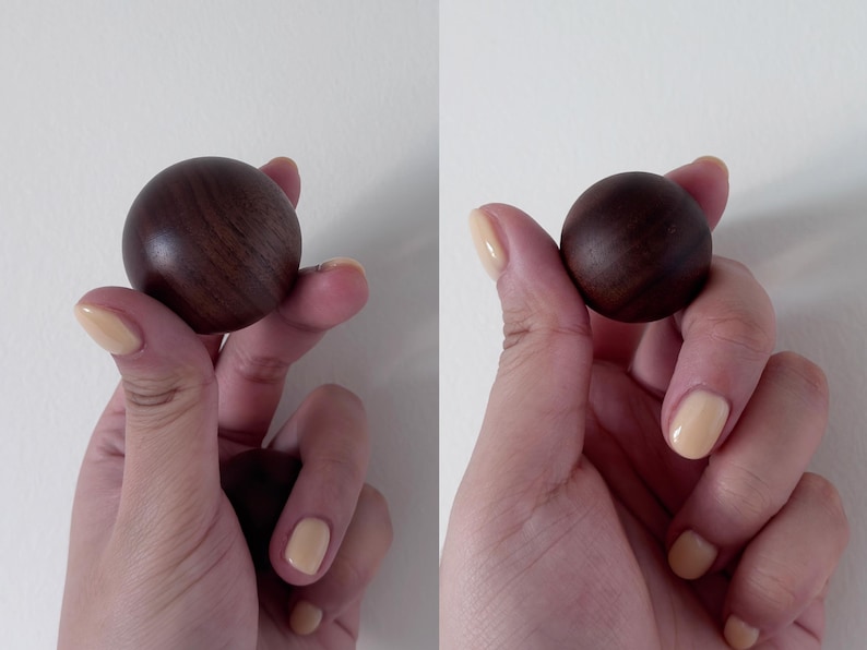 May include: Two dark brown wooden spheres, each held in a hand with light yellow nail polish. The spheres have a smooth, polished finish and a visible wood grain pattern. The background is a plain white wall.