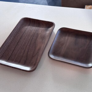 May include: Two wooden trays with a dark brown finish. One is rectangular, and the other is square. Both have rounded edges and a smooth, polished surface. The trays are likely for serving or display.