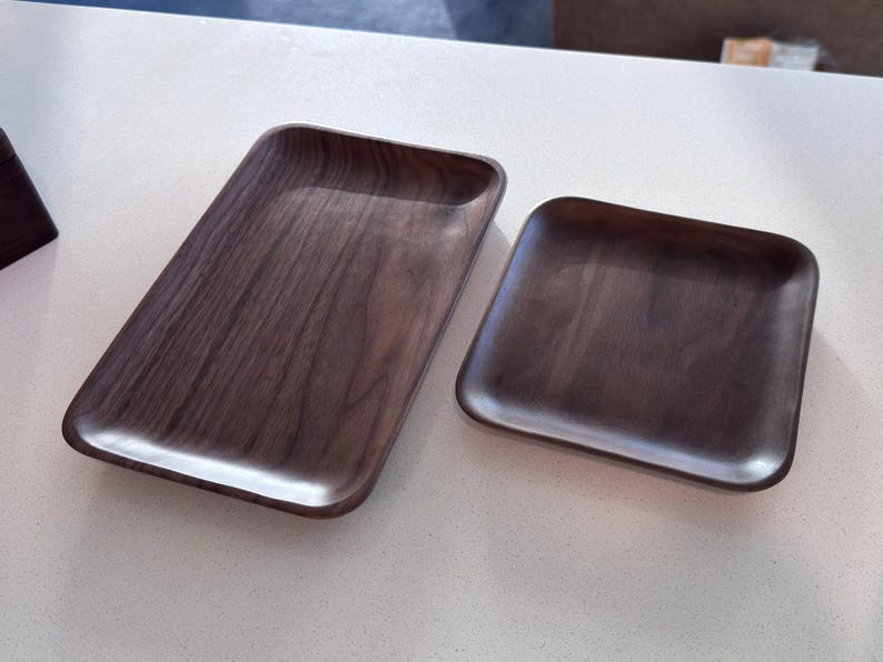 May include: Two wooden trays with a dark brown finish. One is rectangular, and the other is square. Both have rounded edges and a smooth, polished surface. The trays are likely for serving or display.