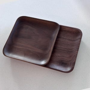 May include: Two square wooden trays with rounded corners are displayed. The trays are made of dark brown wood, showcasing the natural grain. The trays are stacked on top of each other, with one slightly overlapping the other.