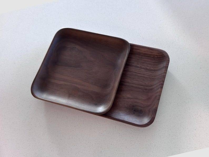 May include: Two square wooden trays with rounded corners are displayed. The trays are made of dark brown wood, showcasing the natural grain. The trays are stacked on top of each other, with one slightly overlapping the other.