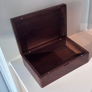 May include: A dark brown wooden box with a hinged lid, shown open. The box is rectangular and has a polished finish. The interior is empty. The box is made of wood, and the lid is held open, revealing the inside.
