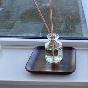 May include: A clear glass diffuser bottle with wooden reeds, sitting on a dark brown square tray. The bottle contains a light-colored liquid. The setup is placed on a white windowsill, with a window in the background.