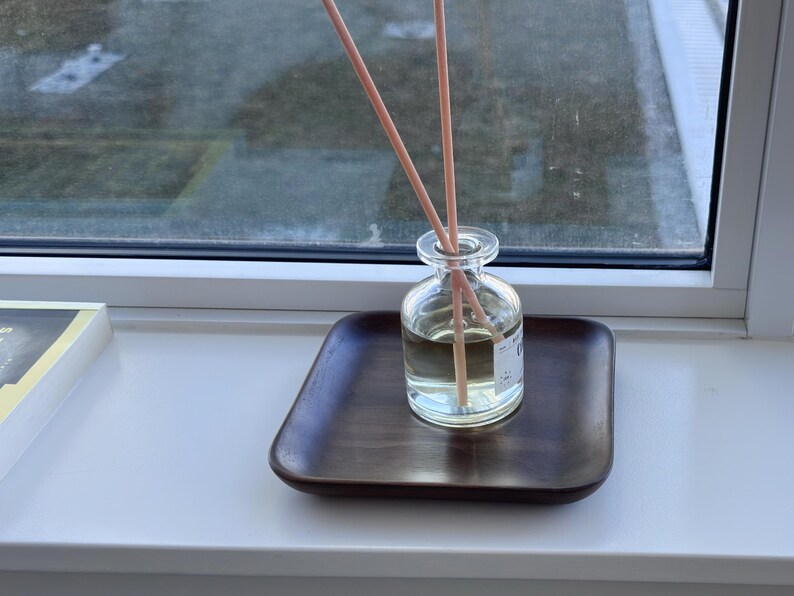May include: A clear glass diffuser bottle with wooden reeds, sitting on a dark brown square tray. The bottle contains a light-colored liquid. The setup is placed on a white windowsill, with a window in the background.