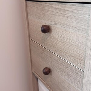 May include: Close-up of a light-coloured wooden cabinet with two drawers. Each drawer has a dark brown, round knob. The wood grain is visible, and the cabinet is set against a light pink wall.