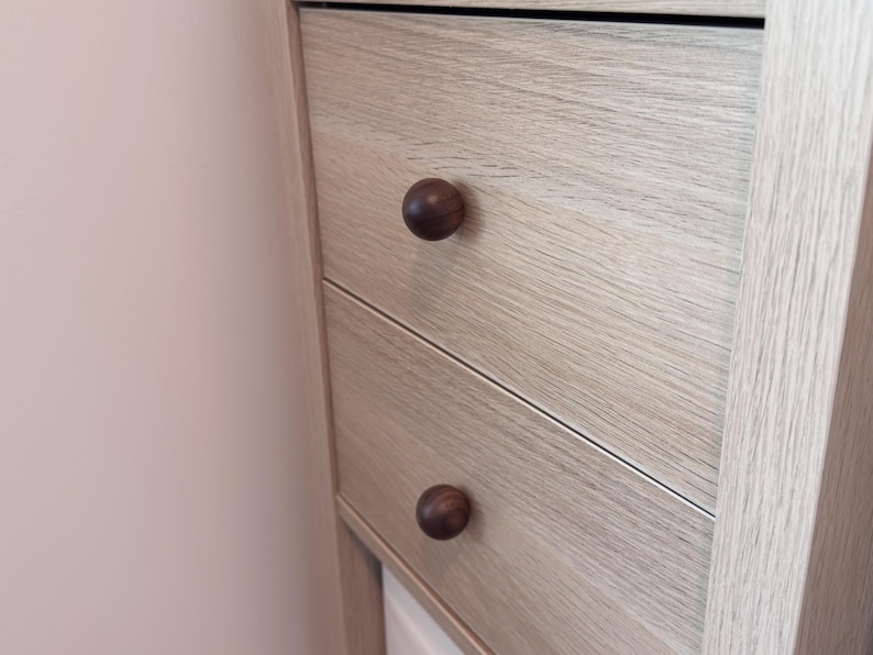May include: Close-up of a light-coloured wooden cabinet with two drawers. Each drawer has a dark brown, round knob. The wood grain is visible, and the cabinet is set against a light pink wall.
