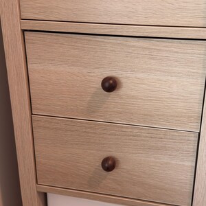 May include: Close-up of a light-coloured wooden cabinet with three drawers. Each drawer has a dark brown, round knob. The cabinet has a simple, modern design with visible wood grain.